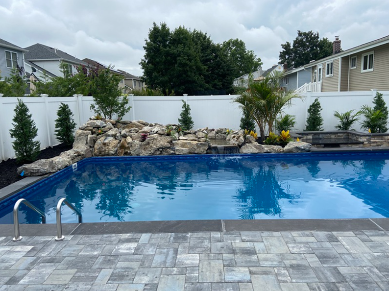 A backyard pool with a rock waterfall and landscaping, surrounded by a white fence and houses.