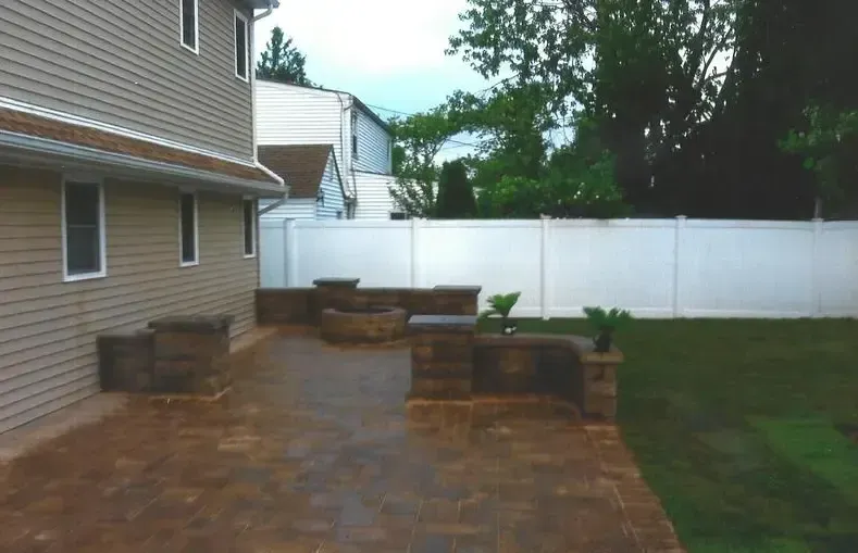 Brick patio with stone columns, a fire pit, and a tan house next to a white fence and green grass.