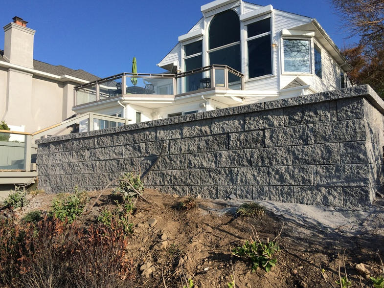 Gray stone retaining wall supports a white house with glass railings on a sunny day.