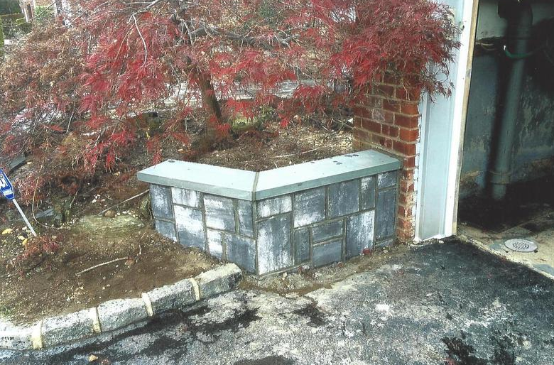 Stone planter with red-leafed tree, next to a brick wall and garage door on asphalt.