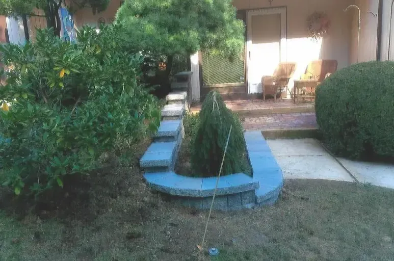 A front yard with a circular blue-bricked planter, trimmed greenery, and a patio with furniture.