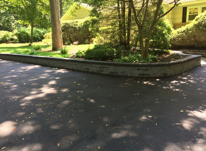 Asphalt driveway with brick retaining wall bordering a garden bed, trees and a yellow house in the background.