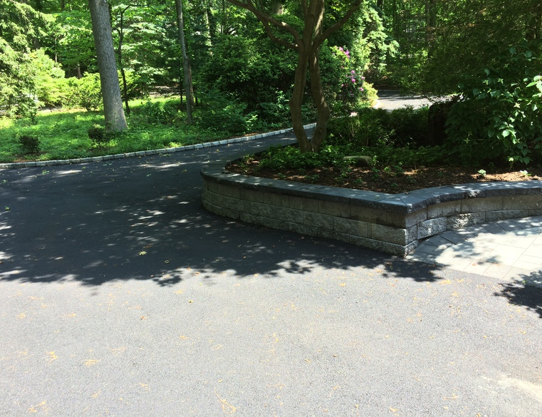 Asphalt driveway curving around a brick retaining wall with a small tree and greenery.