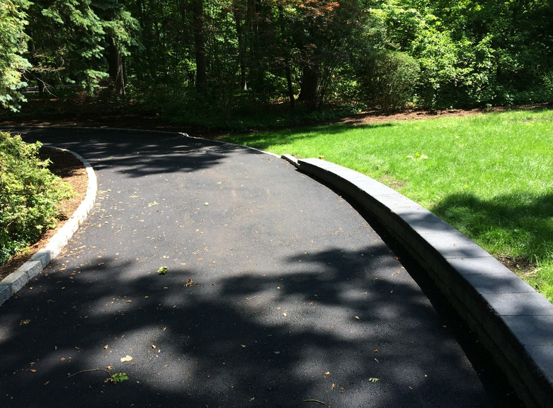 Asphalt driveway curves past a low wall and green lawn, leading toward a wooded area.
