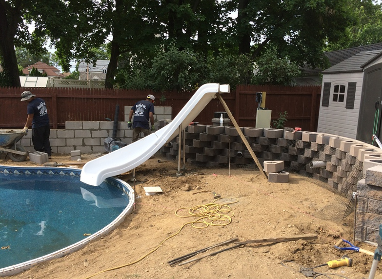 Construction site with workers building a retaining wall around a pool. A white slide is installed.