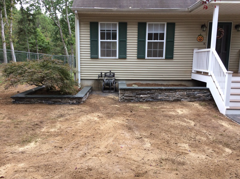 A beige house with dark green shutters, a stone retaining wall, and a small tree, surrounded by dirt.