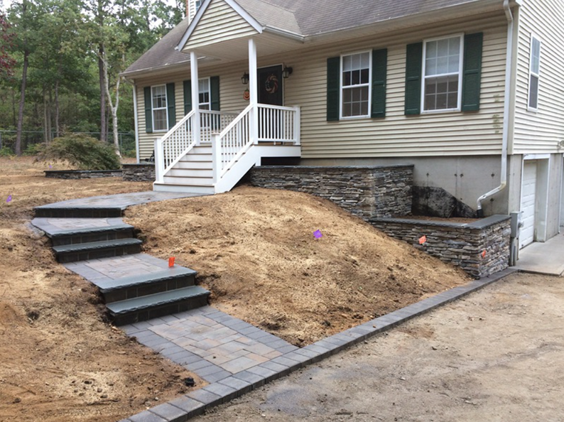 Beige house with green shutters, stone retaining walls, and paved walkway and steps.