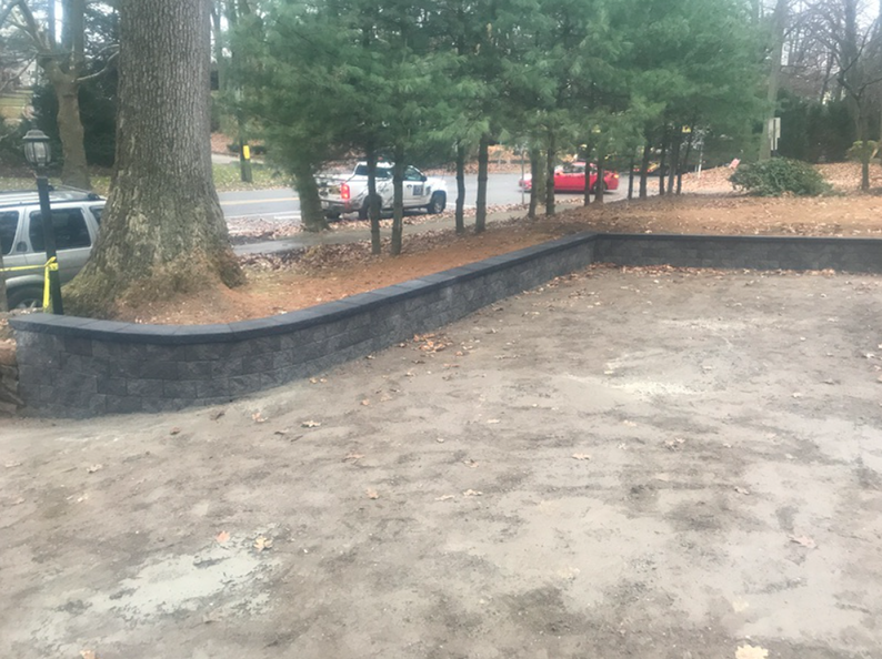 Gray retaining wall bordering a gravel driveway, next to trees and a road.