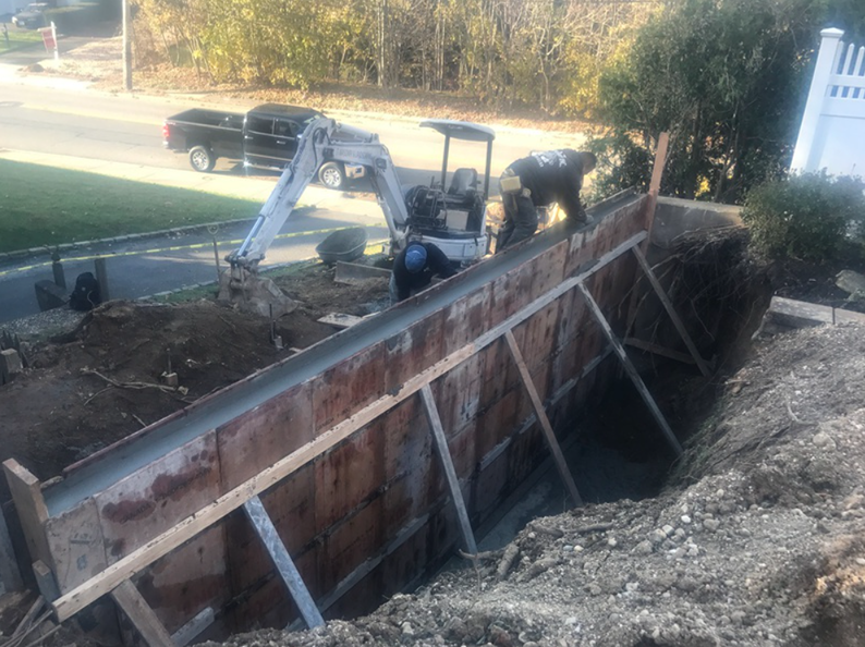 Workers pouring concrete into wooden formwork in an excavated area, excavator and truck in background.
