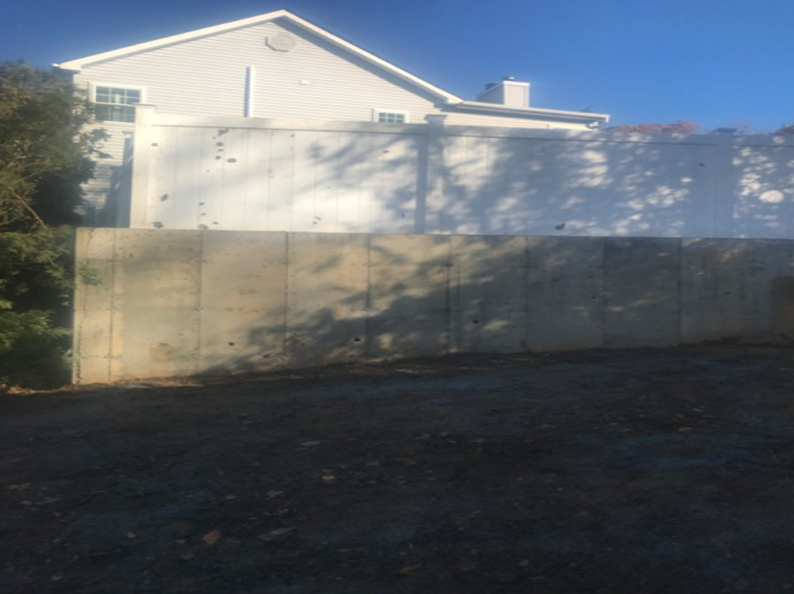 Concrete wall in front of a light-colored building under a blue sky. Shadows on the wall and ground.