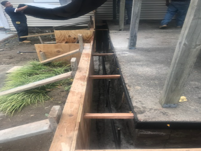 Construction site: wooden forms for concrete trench next to a building, workers in background.