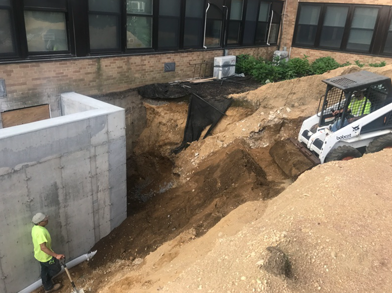 Construction site next to a building. A worker stands by a concrete wall, while a skid steer moves dirt.