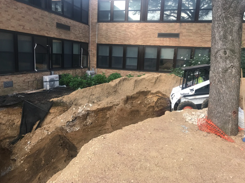 Construction site with a trench being dug near a brick building. A skid steer loader is present.