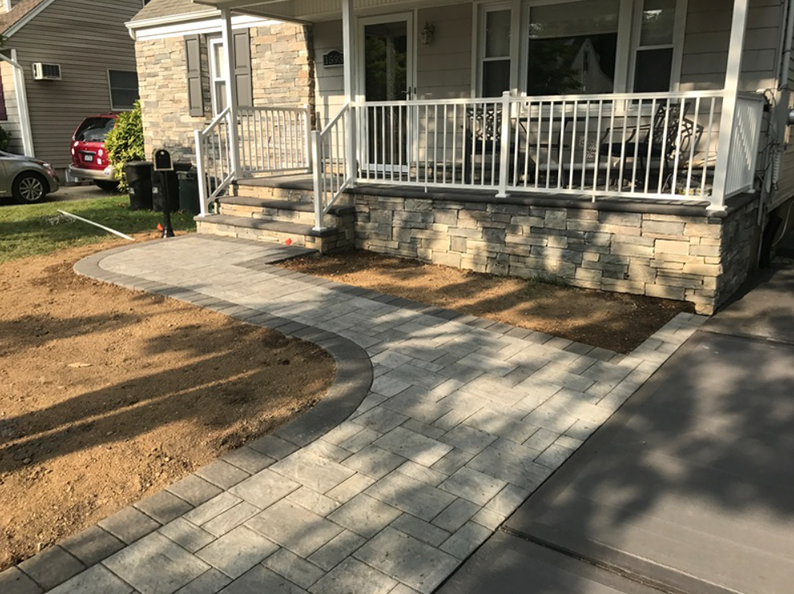 Stone walkway leading to a house with a white porch. Brick pavers are grey with brown dirt.