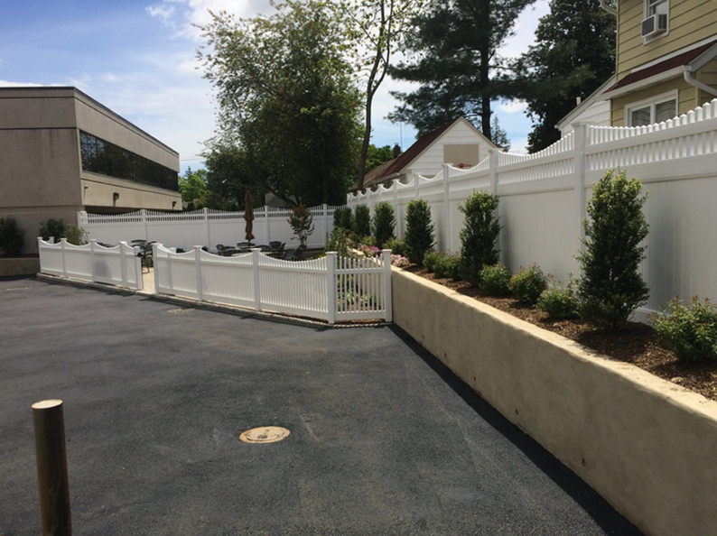 White picket fence surrounds a planted area with bushes, adjacent to a building with asphalt.