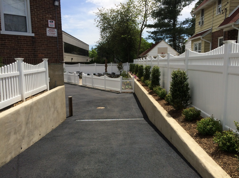Asphalt driveway bordered by white picket fence and retaining wall with small green bushes.