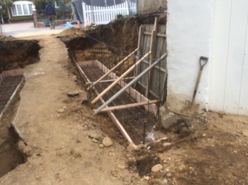 Construction site: Excavated trench with wooden forms for a retaining wall, near a white fence and building.