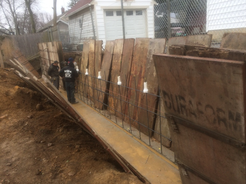 Construction site with wooden forms, rebar, and workers in a narrow trench.