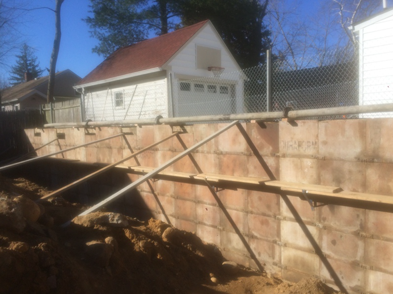 A retaining wall under construction with support beams, dirt, and a small white building with an orange roof.