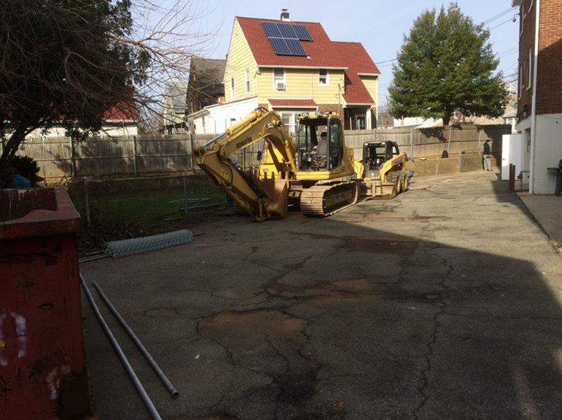 Yellow excavator and loader on a cracked asphalt surface in front of a fenced yard, house in the background.