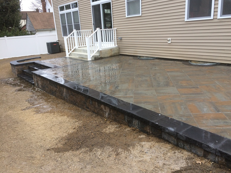 Backyard patio with pavers, retaining wall, stairs leading to a raised deck, and house.