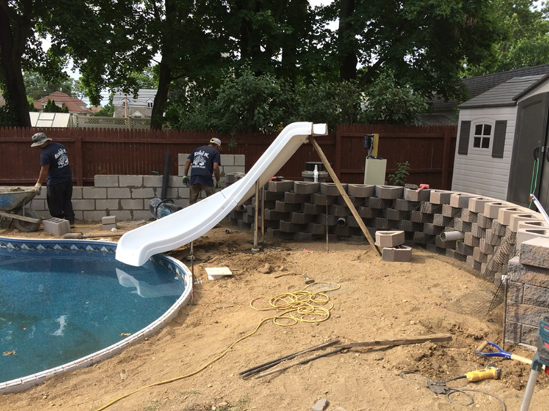 Workers building retaining wall around a pool with a white slide; construction site.