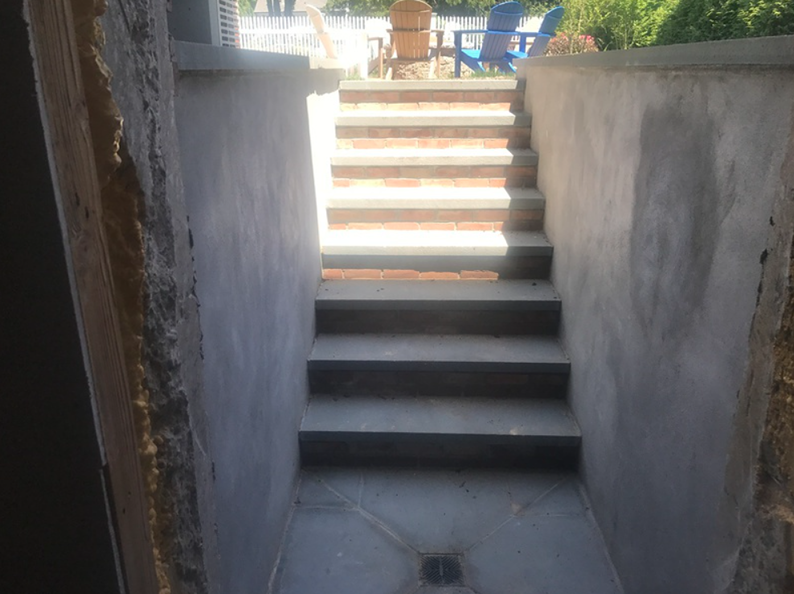 Stone steps leading up from a gray-walled recessed area to an outdoor patio with blue chairs.