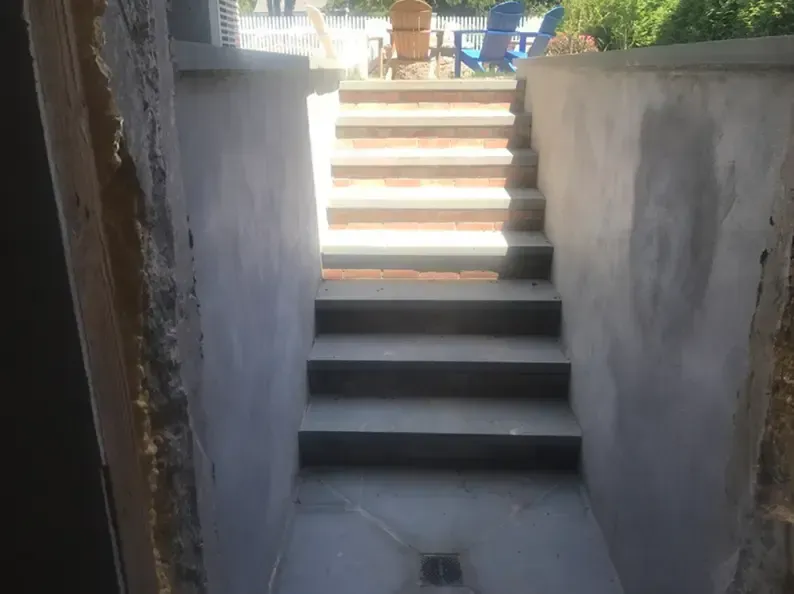 Staircase leading upwards from a recessed area, with concrete steps and walls. Bright sunlight at top.