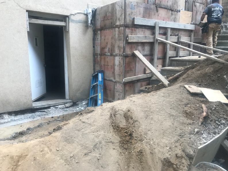 Construction site with exposed dirt, wooden forms, stairs, and a partially open doorway. A worker is present.