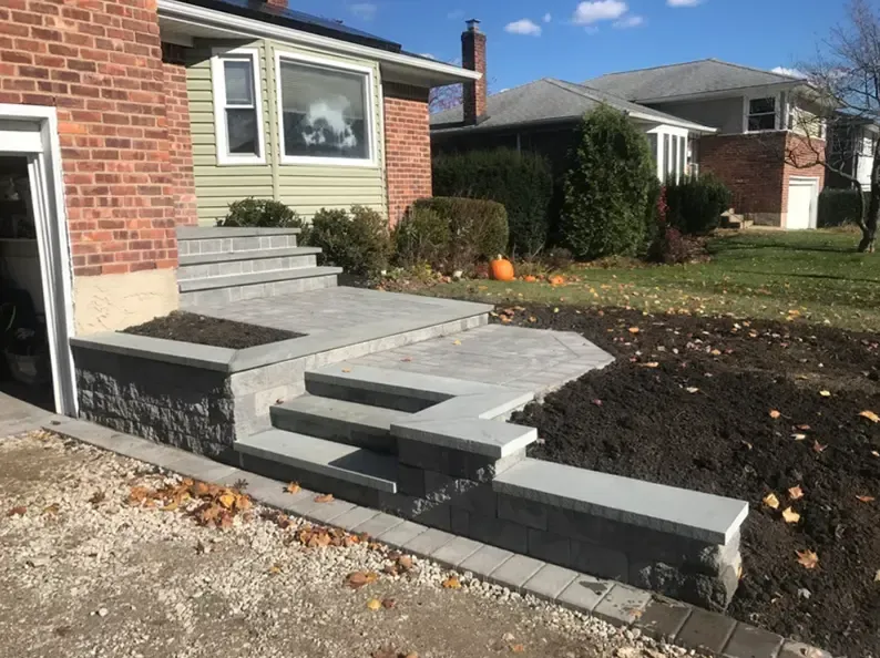 Brick home entrance with gray stone steps, retaining walls, and fresh soil; fall leaves and a pumpkin visible.