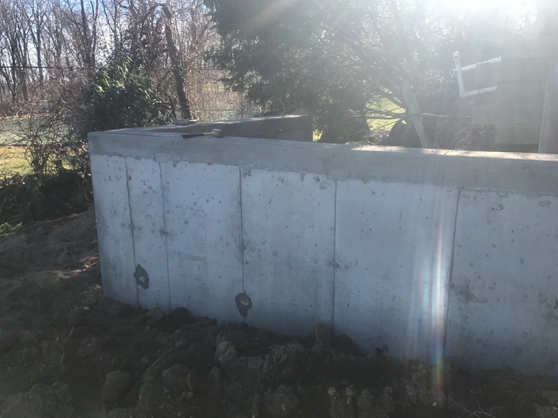 Concrete foundation walls, outdoors, with a grassy foreground and trees in the background under a bright sun.