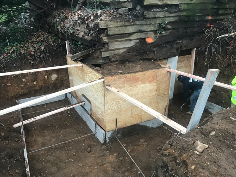 Construction site: wooden forms for a concrete foundation, supporting an old wooden structure, worker in background.