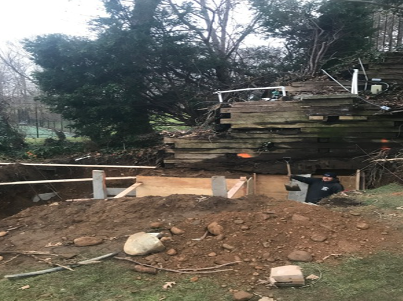 Construction site with a worker, soil, wooden frames, and a retaining wall in a grassy, wooded area.
