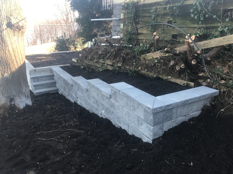 A gray block retaining wall with steps leading up to a raised garden bed, set outside.