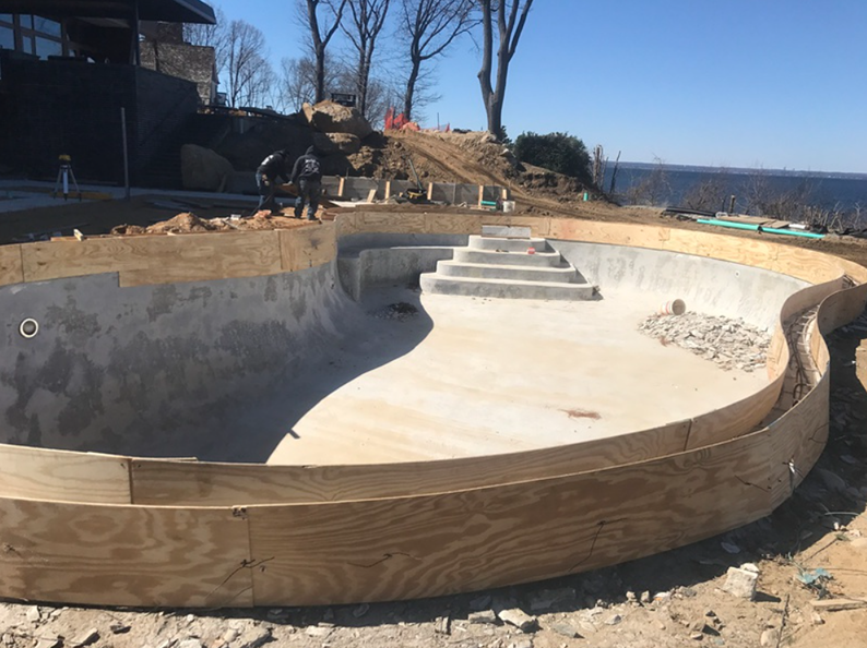 Construction of an in-ground pool, with wooden forms, steps, and workers, overlooking a lake.