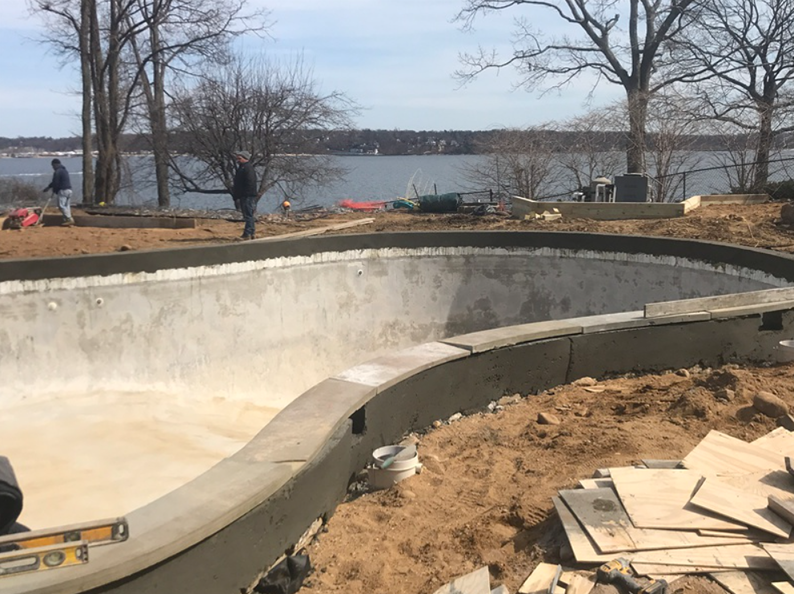 Concrete skate bowl under construction near water, two people in background.