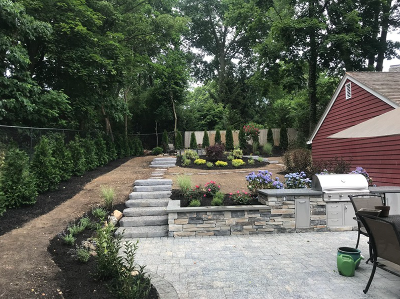 Landscaped backyard with stone patio, grill, steps, and garden beds. Red house and trees in the background.
