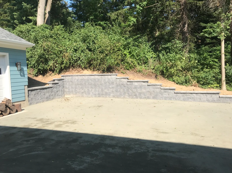 Gray retaining wall next to a paved area, with a wooded hill in the background, beside a light blue building.