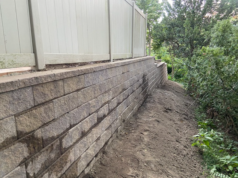 Retaining wall of stacked, brown blocks, supporting a fence and earth, with greenery to the right.