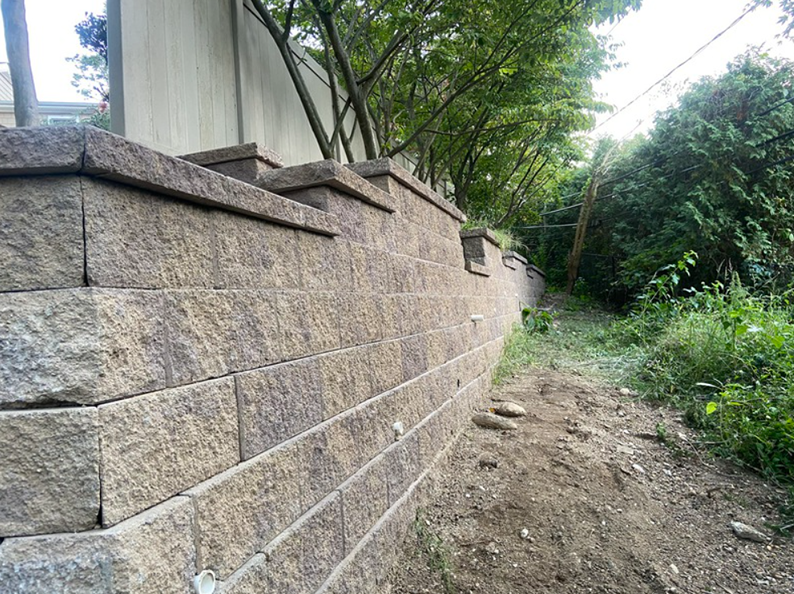 Stone retaining wall with capstones, along a sloped embankment, trees in the background.