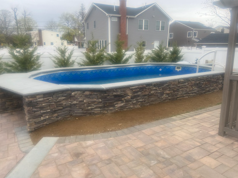 Stone-walled pool with blue interior, surrounded by a paved patio and evergreens, with houses in the background.