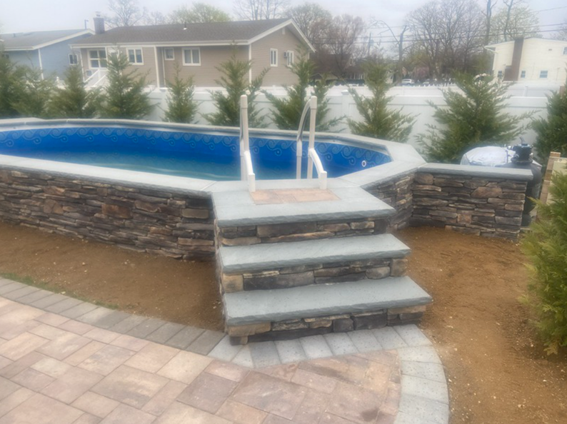 Stone-faced pool with steps and a ladder. Brown and gray stone, with trees and a house in the background.
