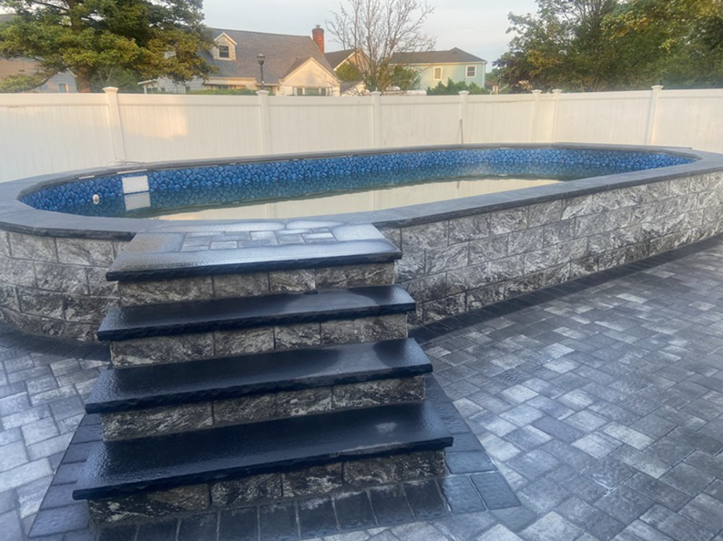 Oval-shaped pool with gray stone wall and steps; paved patio and white fence in background.