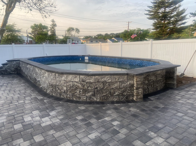 Oval-shaped in-ground pool with stone retaining walls. Paved patio, white fence, and houses in the background.