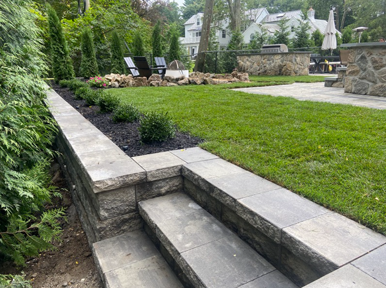 Stone steps leading up to a raised garden bed with green grass, shrubs, and a fire pit.
