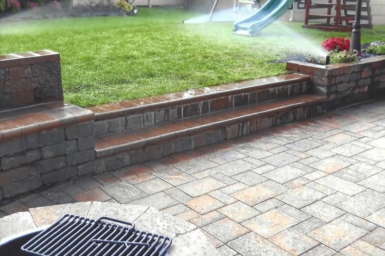 Stone patio with brick steps leading to a grassy lawn with sprinkler, a grill in the foreground.