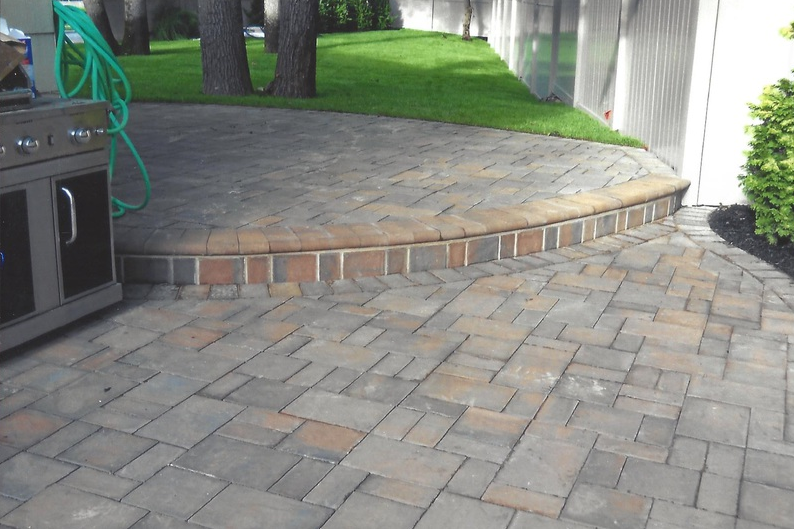 Paver patio with curved edge, leading to a grassy area. A grill and green hose are visible.