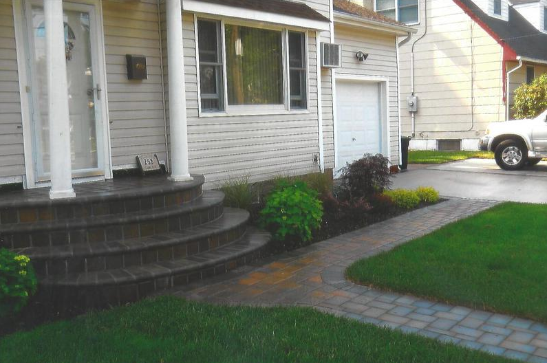 Brick pathway leads to home with stone steps, flanked by green grass and a small garden.