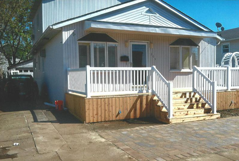 House with white railing, wooden steps, and a paved driveway on a sunny day.