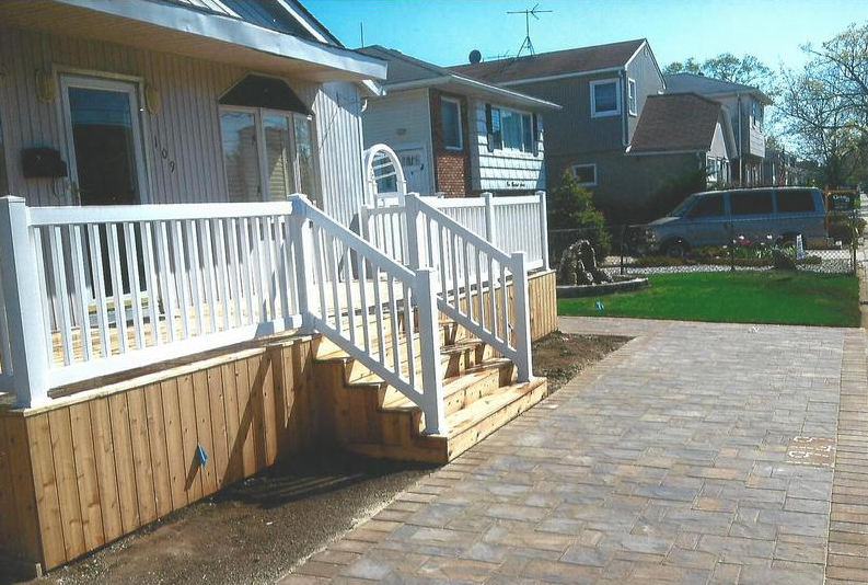 White-railed wooden deck and steps leading to a house, brick paved walkway.